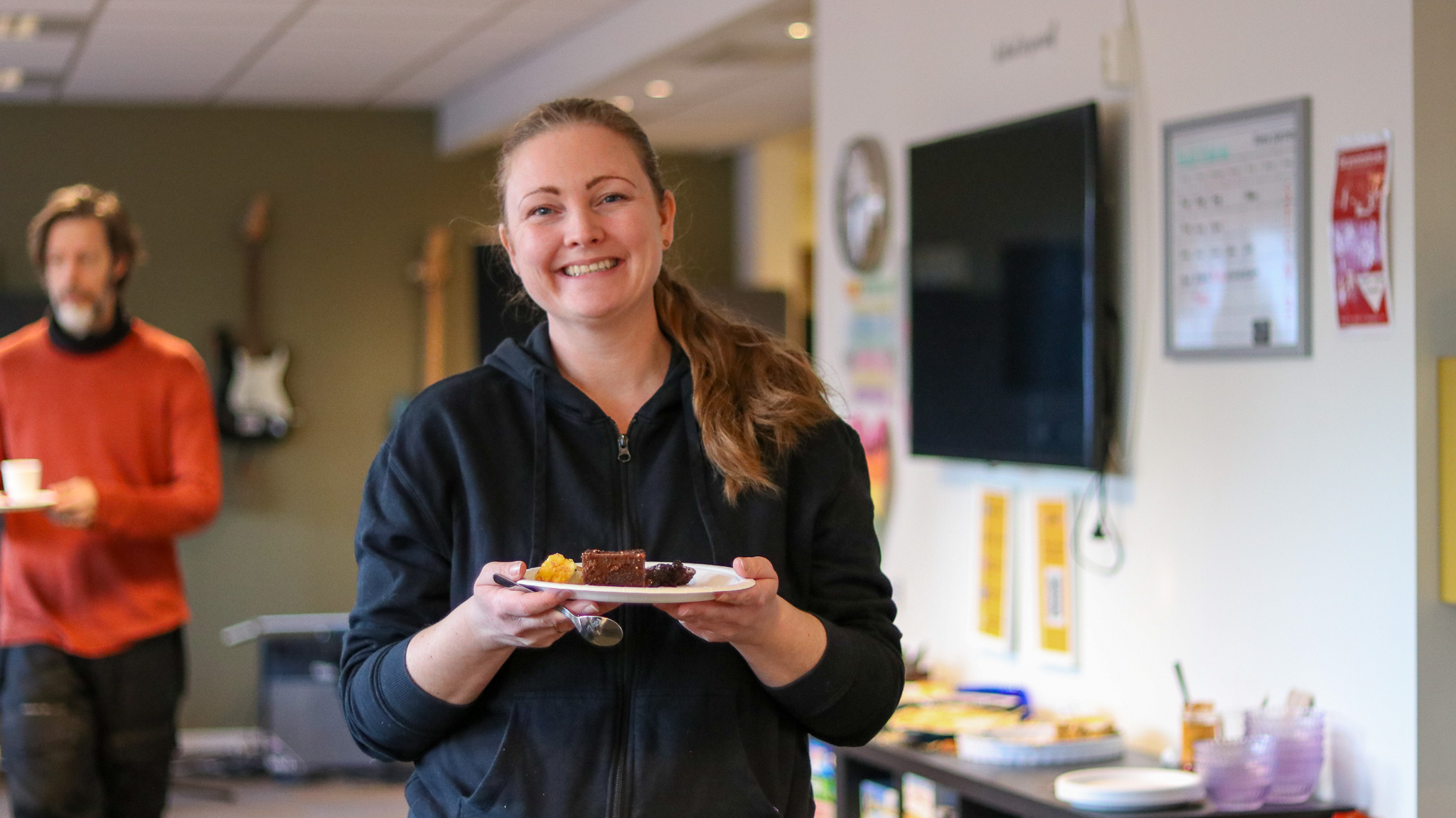 Bilde av en smilende dame som holder en tallerken med kake på. I bakgrunnen ser vi flere kaker og en person som holder en tallerken.