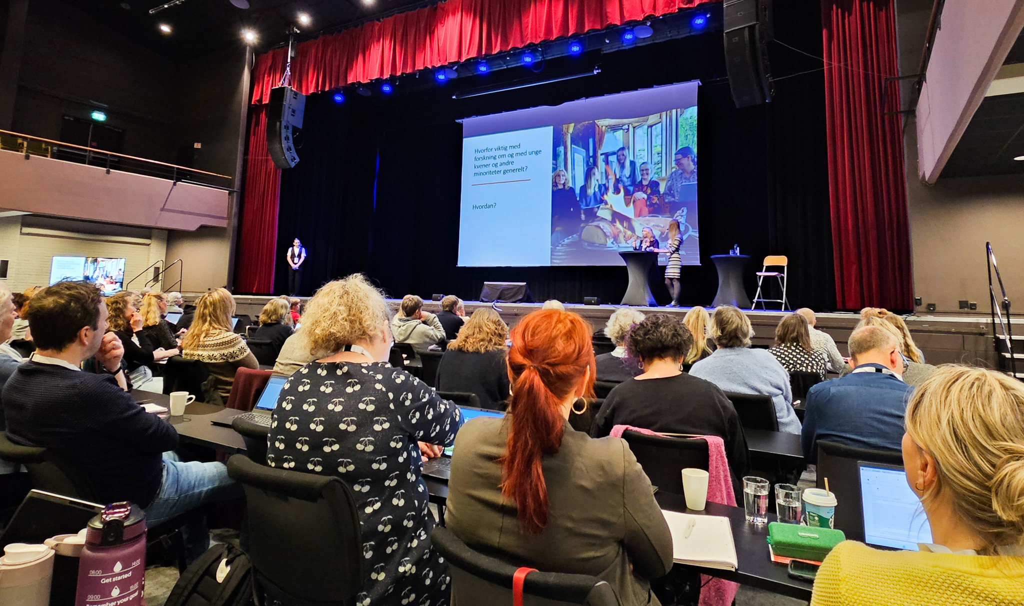 Stemningsbilde fra konferanse. Publikum sitter på rekker. På scenen står tre personer. Det er rødt sceneteppe og et stort lerret som viser bilde av mennesker rundt et bål.